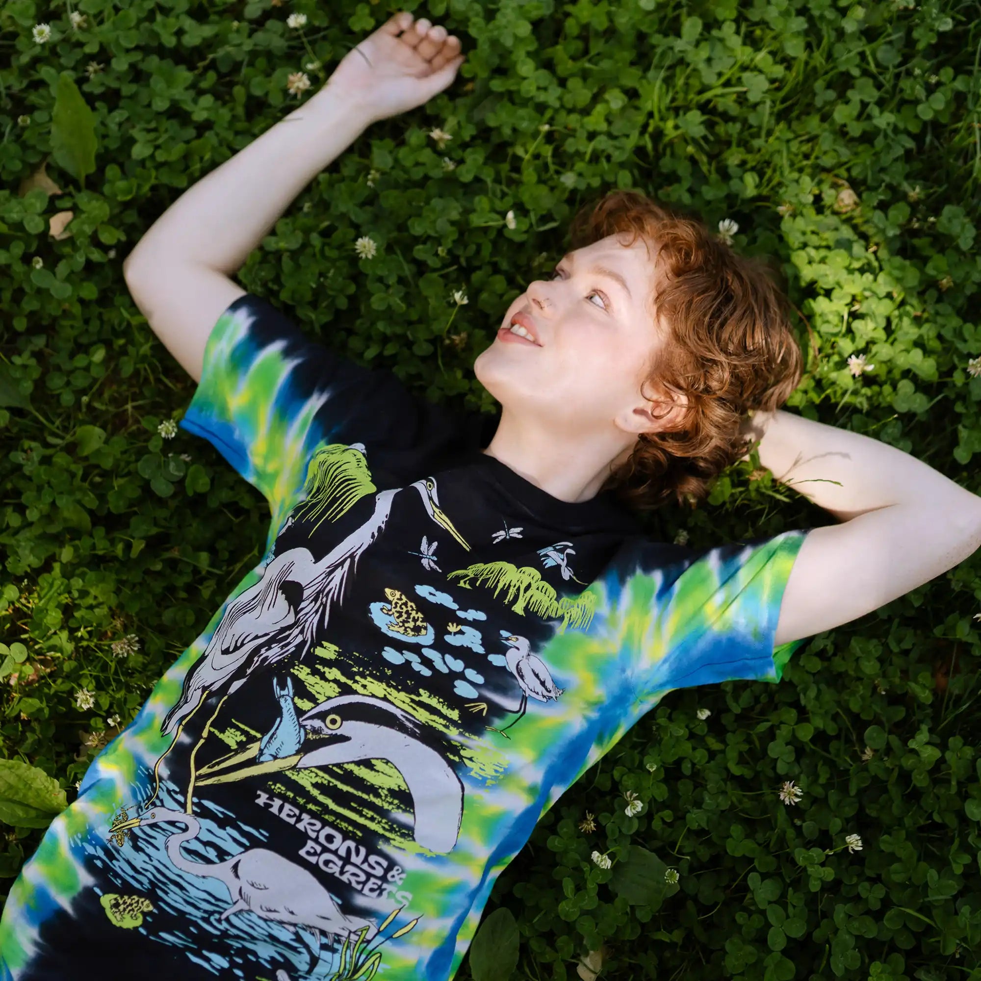 Colorful lagoon t-shirt with a nature-themed design, lying on grass.