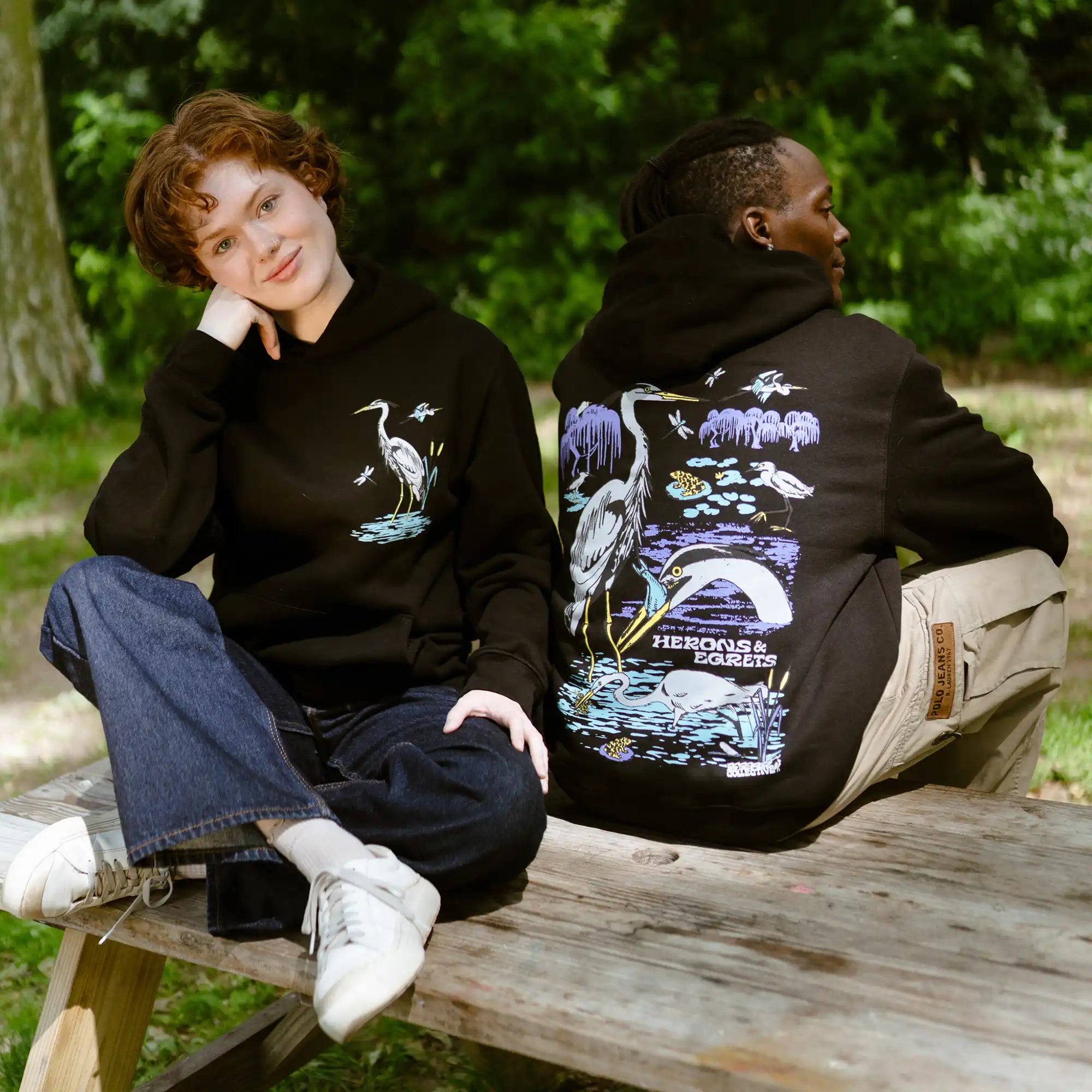 Two people wearing black hoodies with nature-themed designs sitting on a wooden bench outdoors.