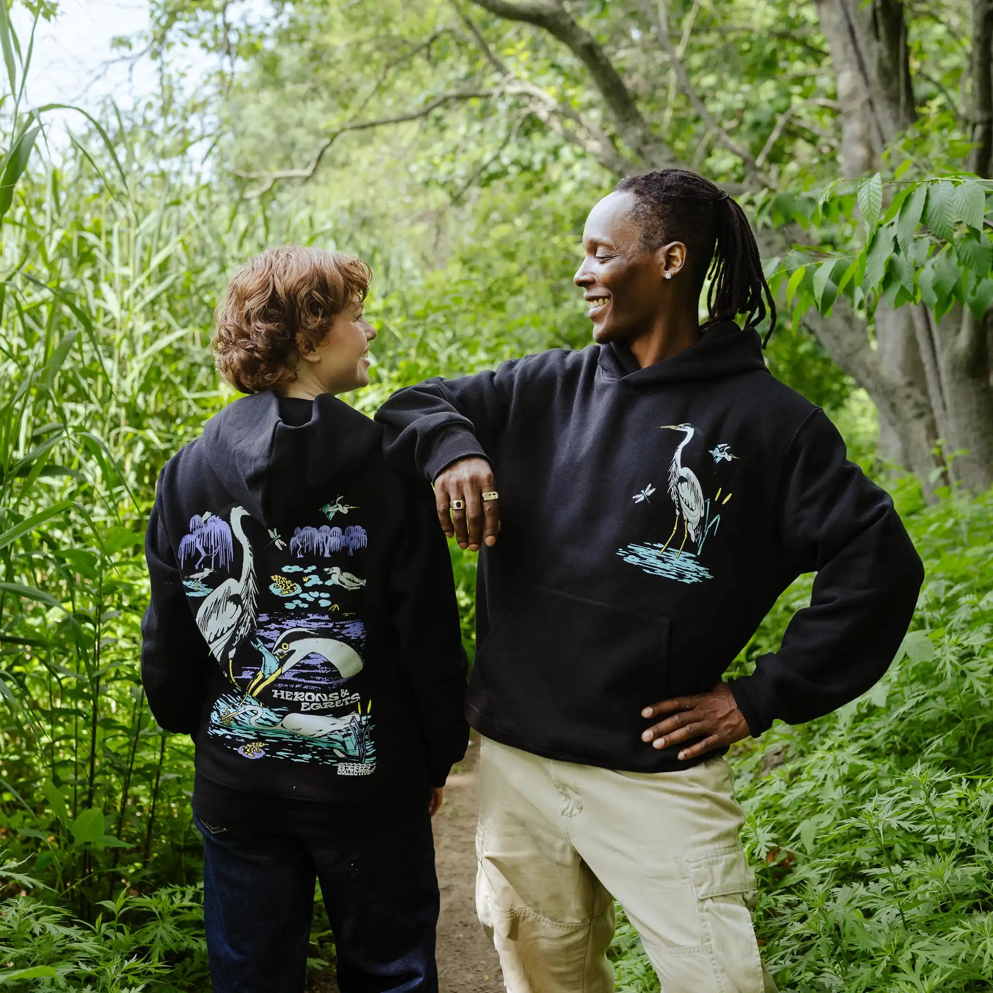 Two people wearing black hoodies with nature-themed designs in a forest setting