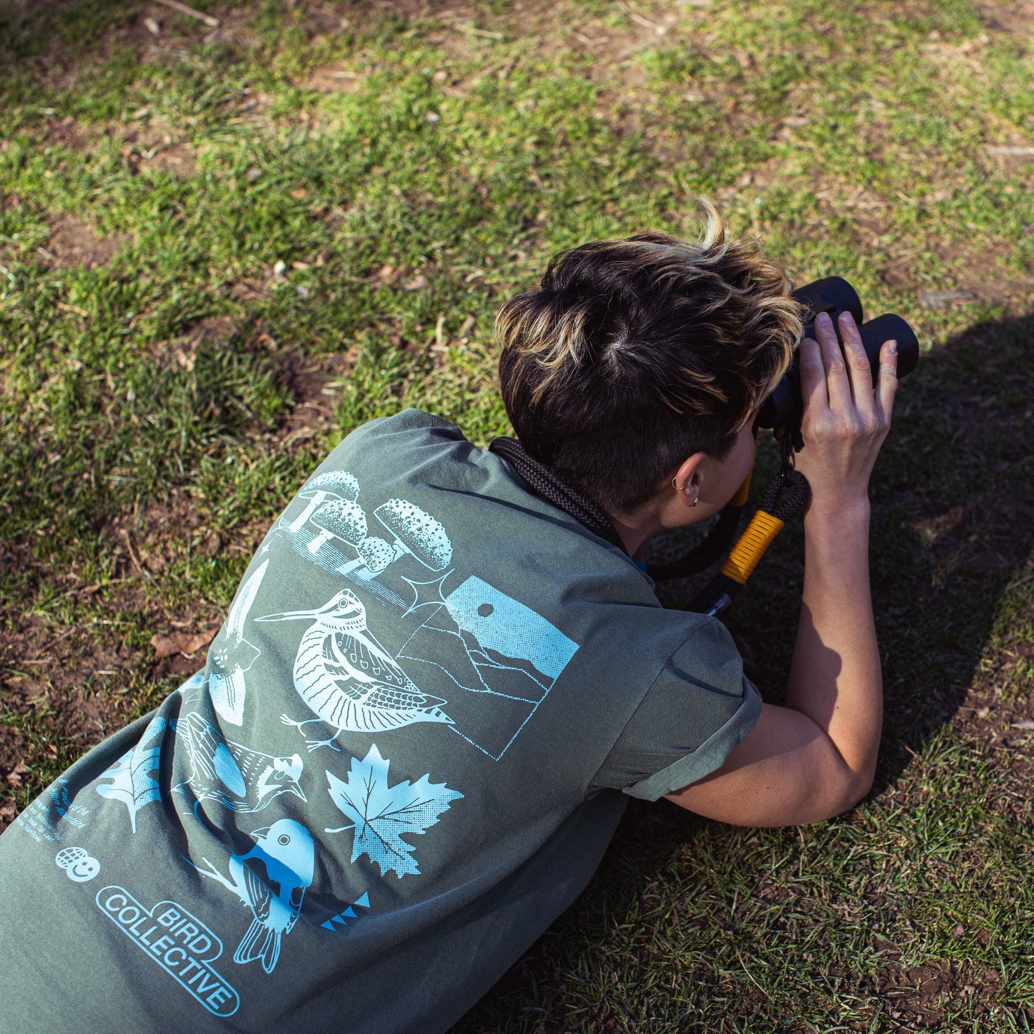 Photo of a birder laying on the grass, looking through binoculars and wearing a Bird Collective x American Bird Conservancy Appalachian Woodland T-Shirt